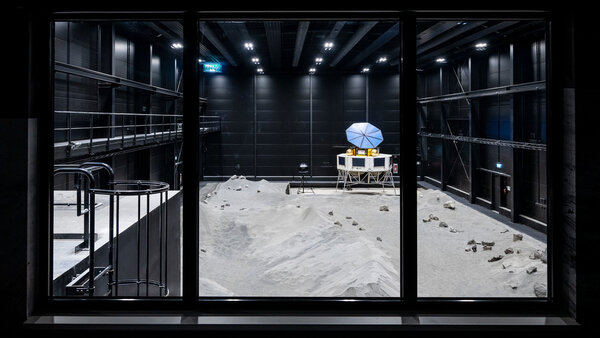 A lunar simulation room with a sandy landscape, rocks, and a structure with a blue umbrella-like device inside a black-walled facility.