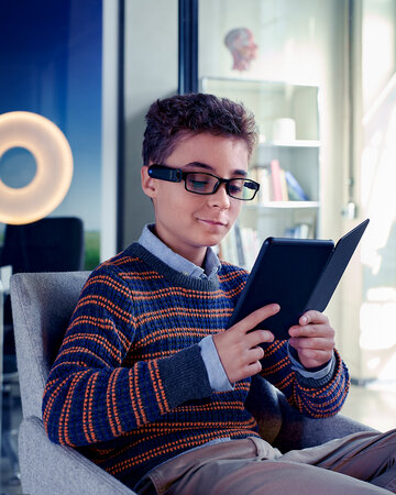 Ein Junge mit Lesebrille mit integrierter digitaler Kamera sitzt in einem modernen Raum und hält ein Tablet in den Händen.