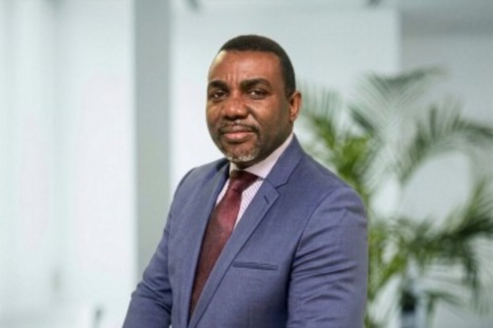 A portrait photograph of a man in a suit and tie standing in an office environment with plants in the background.