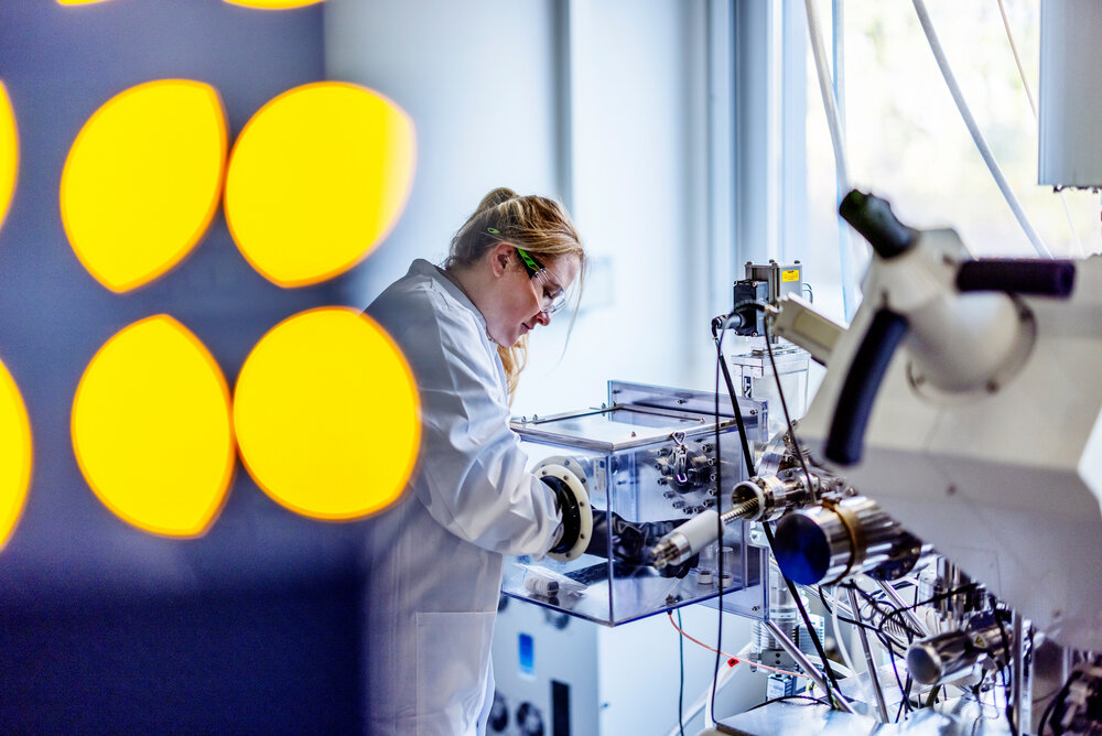 A woman in a white lab coat is working on a laboratory instrument while wearing protective gloves.