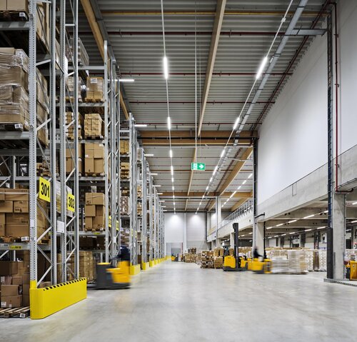 Großes Logistiklager mit hohen Regalen voller Kartons, gelbe Stapler bewegen Güter in den breiten Gängen.