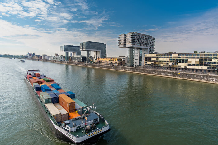 Ein Containerschiff auf dem Rhein mit bunten Containern, im Hintergrund Kölner Kranhäuser im Rheinauhafen und Uferbebauung.