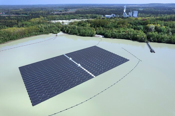 Eine schwimmende Solaranlage auf einem See, umgeben von einem Wald und mit einem Kraftwerk im Hintergrund.