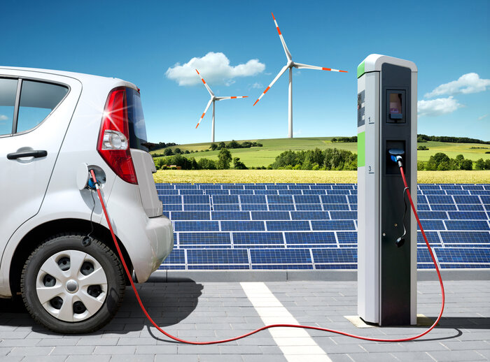 Ein Elektroauto lädt an Ladestation, im Hintergrund Solarpanels und zwei Windräder auf einem grünen Feld unter blauem Himmel.